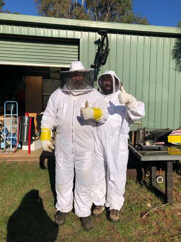 Duas pessoas com roupas e luvas brancas de apicultor estão do lado de fora de um galpão, sorrindo e fazendo sinal de positivo, prontas para as atividades de apicultura em um dia ensolarado.
