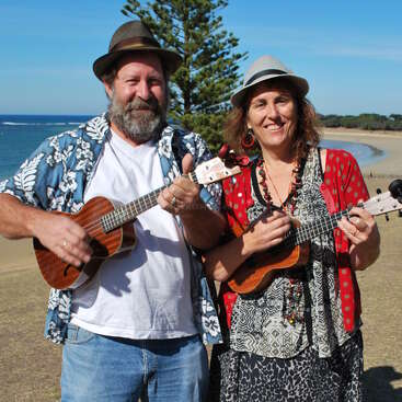 A imagem mostra um homem e uma mulher lado a lado em uma praia, cada um segurando um ukulele e usando chapéus. Eles estão vestidos com roupas casuais.
