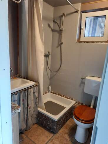 This is a small bathroom with a shower area, a curtain, a window, a toilet with a wooden seat, and a sink with floral curtains.