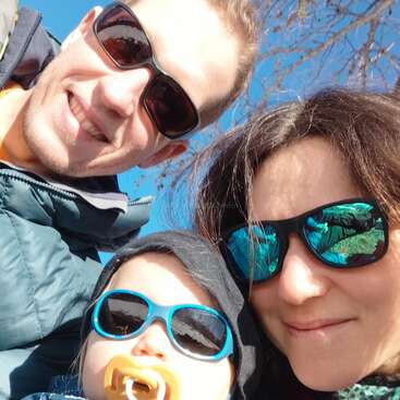 The image shows a family of three, with a man, woman, and baby, all wearing sunglasses, posing for a selfie on a sunny day.