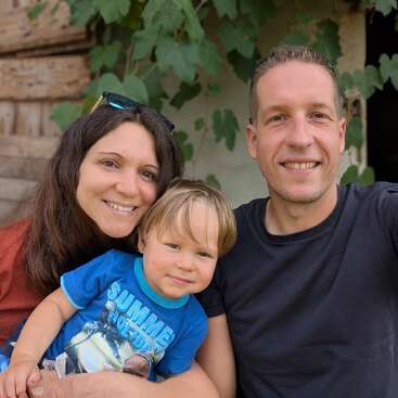Eine glückliche dreiköpfige Familie posiert gemeinsam im Freien und lächelt herzlich. Grüne Blätter und rustikale Holzbretter bilden einen gemütlichen, natürlichen Hintergrund. Das Kind trägt ein blaues Hemd.