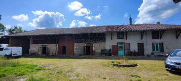 Un rústico edificio rural con tejado de tejas se alza bajo un cielo azul y brillante. Dos vehículos, hierba verde y algunos muebles de exterior completan la apacible escena.