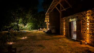 Un rústico granero de piedra se ilumina suavemente por la noche. Las cuerdas de luces y los farolillos crean un ambiente acogedor. La gente está reunida fuera, disfrutando de una noche tranquila.