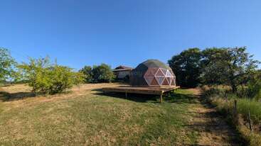 Una estructura de cúpula geodésica se asienta sobre una plataforma de madera en un campo cubierto de hierba, rodeada de árboles y arbustos, bajo un cielo azul claro y brillante.