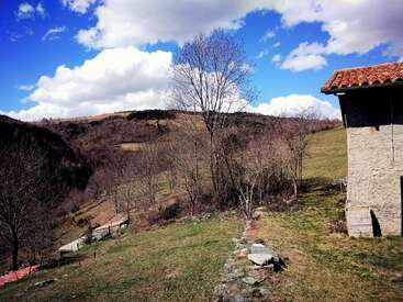 La imagen representa un sereno paisaje rural con un edificio de piedra con tejado de tejas, sobre un fondo de colinas onduladas y un cielo azul con nubes blancas.