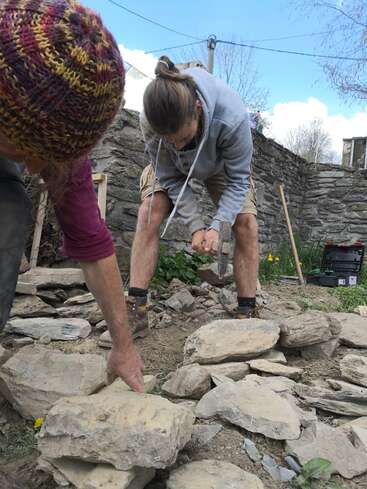 Dos personas trabajan en un muro de piedra, utilizando herramientas para colocar piedras planas, con un muro de piedra y un cielo azul de fondo.