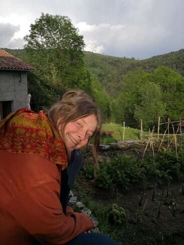 La imagen muestra a una mujer de pelo largo y chaqueta roja, agazapada en un jardín, rodeada de exuberante vegetación y un paisaje sereno bajo un cielo nublado.