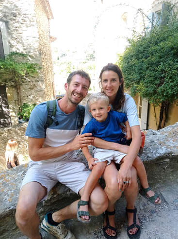 Uma família alegre de três pessoas — pai, mãe e filho pequeno — senta-se ao ar livre em um muro de pedra, cercada por vegetação e prédios rústicos de pedra, sorrindo feliz.