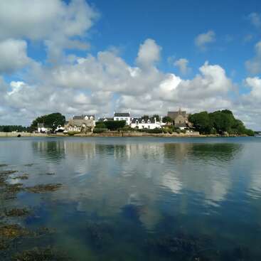 A peaceful village with white houses and a church sits by a calm body of water, reflecting clouds and blue sky, surrounded by greenery and trees.