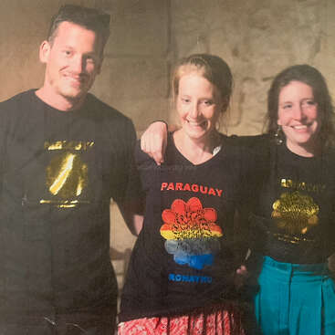 Three smiling people stand together, arm in arm, wearing black shirts. The center shirt features "PARAGUAY ROHAYHU" with a colorful flower design, conveying friendship.