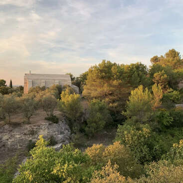 The image depicts a serene landscape featuring a white house nestled among trees and bushes on a hillside, with a cloudy sky above.