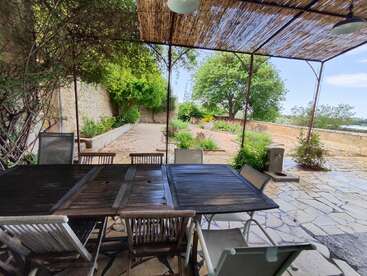 A rustic outdoor patio with a wooden table and chairs under a bamboo canopy, overlooking a stone-paved garden surrounded by greenery and a stone wall. Peaceful atmosphere.