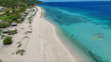 Une plage tropicale immaculée avec du sable blanc, des eaux turquoise cristallines, une végétation verte éparse et des huttes ombragées le long du rivage. Des collines verdoyantes s'élèvent doucement à l'arrière-plan.