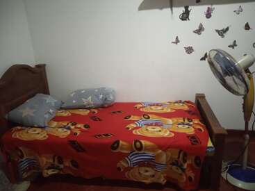 L'image représente une petite chambre confortable avec un lit simple, orné d'une couverture rouge avec des ours en peluche et d'une tête de lit marron, accompagné d'un ventilateur et de papillons sur le mur.