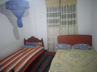 L'image représente une petite chambre à coucher avec deux lits, l'un avec un cadre en bois et l'autre avec une tête de lit rouge, tous deux ornés d'une literie et de rideaux à motifs.