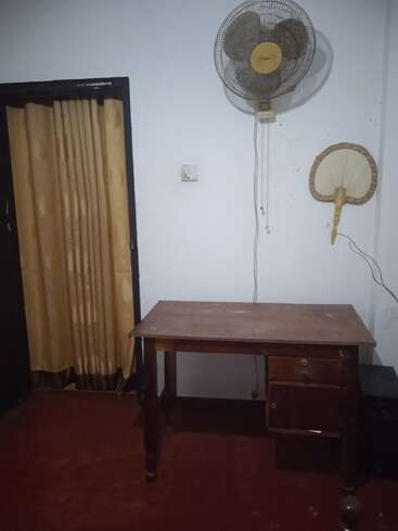 L'image représente une pièce avec un bureau en bois, un ventilateur et un rideau, aux murs blancs et au sol rouge, avec une petite table et un ventilateur au mur.