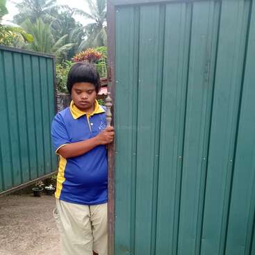 L'image représente un homme debout devant une clôture verte, vêtu d'une chemise bleue et jaune, d'un pantalon kaki et sans chaussures, sur un fond tropical serein.