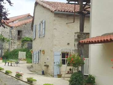 Ein charmantes, rustikales Haus mit Steinmauern, blauen Fensterläden, roten Ziegeldächern, Blumentöpfen und einem kleinen Sitzbereich im Freien in einem ruhigen Innenhof.