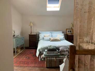 Dieses gemütliche Schlafzimmer hat ein Dachfenster, hellblaue Bettwäsche, Vintage-Möbel, zwei Lampen, einen rot gemusterten Teppich und gefaltete Wäsche, die auf einer Truhe gestapelt ist.