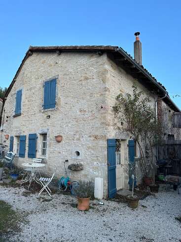 This image shows a charming stone cottage with blue shutters, a tiled roof, and an outdoor seating area, surrounded by potted plants and gravel ground.