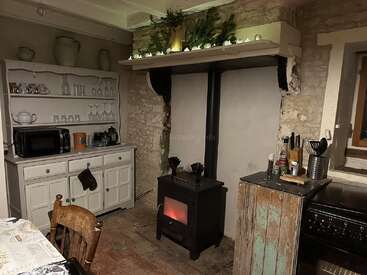 This cozy rustic kitchen features a wood stove, exposed stone walls, vintage hutch with glassware, microwave, wooden furniture, and decorative greenery above the mantel.