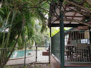 La imagen muestra un entorno sereno al aire libre, con una piscina, muebles de jardín y frondosos árboles, con una valla y una puerta que conducen a un tranquilo oasis en el patio trasero.
