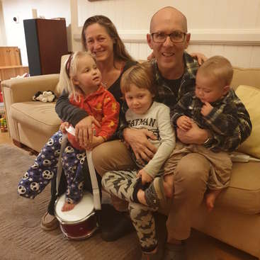 La imagen muestra a una familia de cinco miembros sentados en un sofá color canela en un salón, con una mujer, un hombre y tres niños posando para una foto.
