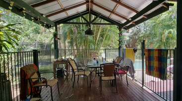 Una terraza cubierta de madera con una mesa de cristal y varias sillas, rodeada por una barandilla negra. Las toallas cuelgan de la valla y una exuberante vegetación rodea la zona.