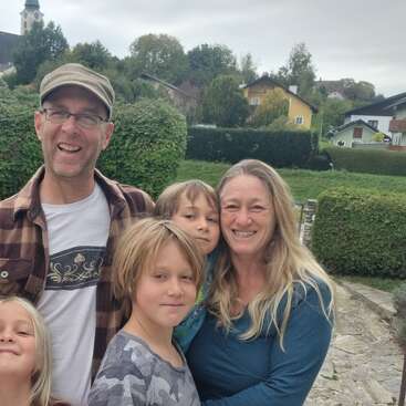 A happy family of five smiles outdoors. Behind them, green hedges, colorful houses, and a church tower create a peaceful, scenic background on a cloudy day.