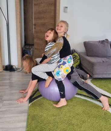 Three laughing children balance together on a large purple exercise ball in a living room, having fun and playing, surrounded by cozy furniture and a green rug.