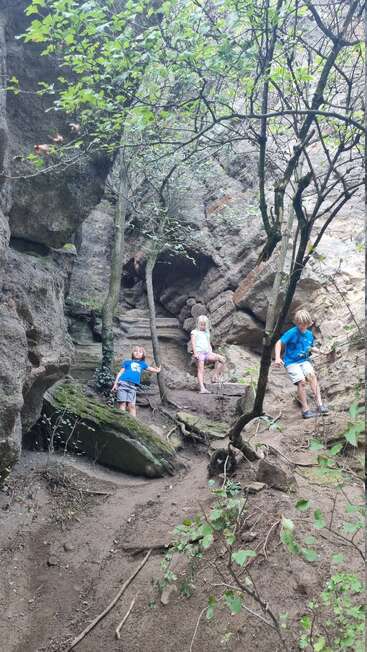 Three children explore a rocky, forested area, climbing among boulders, tree roots, and steep terrain. The setting is lush, rugged, and adventurous, with natural beauty.