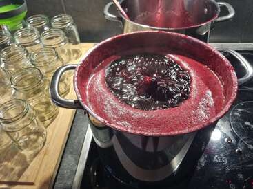 A pot filled with bubbling, dark red jam is boiling on a stove. Empty glass jars for canning are neatly arranged on a wooden board nearby.