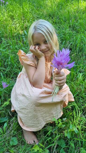 A young girl in a peach dress sits barefoot on grass, holding a bouquet of purple flowers, smiling softly, surrounded by lush green nature and sunshine.