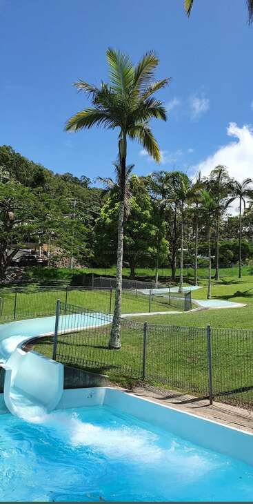 A bright, sunny day at a water park features a curving blue water slide ending in a pool, surrounded by green grass, palm trees, and clear skies.