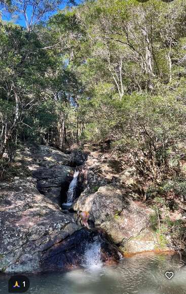 A person in orange swimwear stands on rocks by a small waterfall in a forest, surrounded by lush green trees under a clear blue sky. Peaceful nature.