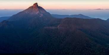 A majestic mountain peak bathed in soft sunrise light rises above dense, forested hills. Mist and distant ridges create a serene and tranquil landscape.
