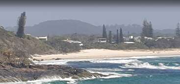 This image shows a beautiful coastline with white sandy beach, turquoise ocean waves, rocky shore, lush greenery, scattered houses, and tall trees under a hazy sky.