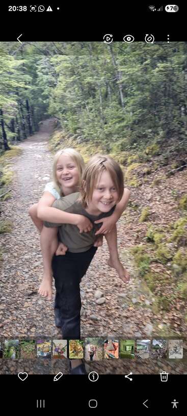 Two children happily walk along a forest trail. One child gives the other a piggyback ride, both smiling joyfully, surrounded by lush, green trees.