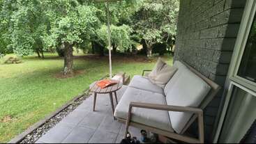 A cozy patio with a cushioned outdoor sofa, small round table, pillows, and open grassy yard. Surrounded by lush green trees, it looks peaceful and inviting.
