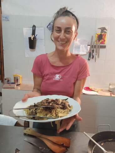 A smiling woman in a kitchen proudly presents a plate of pasta topped with mushrooms. Utensils and kitchen tools are visible on the wall behind her.