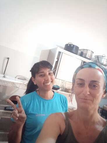 Two women smile cheerfully in a kitchen. One wears a blue shirt and flashes a peace sign; the other has a colorful headband. Pots sit on shelves.