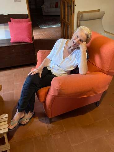 An elderly woman with short gray hair sits comfortably in an orange armchair, smiling warmly, wearing a white shirt, dark pants, and flip-flops in a cozy room.