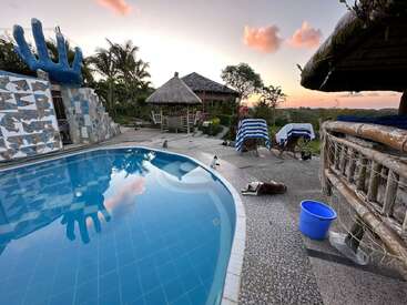 Uma cena tranquila à beira da piscina ao pôr do sol, com espreguiçadeiras com toalhas listradas, uma escultura de mão única, cabanas tiki, palmeiras, um balde azul e cachorros relaxando.