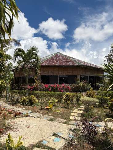 Uma casa rústica com telhado de zinco enferrujado fica em meio a uma vegetação tropical exuberante, flores coloridas e um céu azul cristalino. Um caminho de pedras atravessa o jardim.