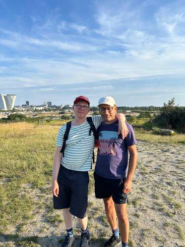 Deux hommes se tiennent côte à côte dehors par une journée ensoleillée, tout sourire. L'un porte une casquette rouge et une chemise rayée, l'autre une casquette blanche et un t-shirt à motif.