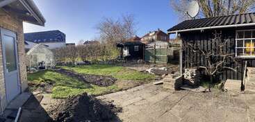 Scène ensoleillée dans un jardin avec une maison en briques, une serre, des plates-bandes, un abri de jardin en bois noir, des pavés empilés, des tas de terre, ainsi que des arbres, des buissons et des maisons voisines.