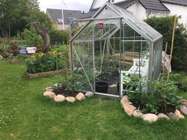 Une petite serre en verre se dresse dans un jardin à l'arrière d'une maison, entourée de plates-bandes surélevées, de plantes verdoyantes, de rochers et d'une pelouse près des maisons.
