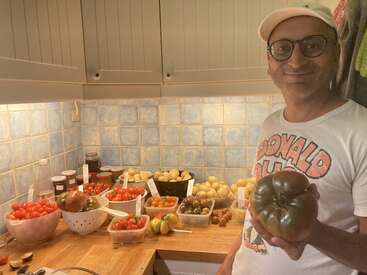Une personne souriante se tient dans une cuisine, tenant une grosse tomate noire. Sur le plan de travail sont disposés divers types de tomates, des pommes de terre, des bocaux et des petits récipients étiquetés.