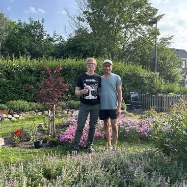 Dois homens estão juntos em um jardim colorido, cheio de flores e vegetação. Eles parecem felizes, aproveitando um dia ensolarado, rodeados por plantas e árvores exuberantes.