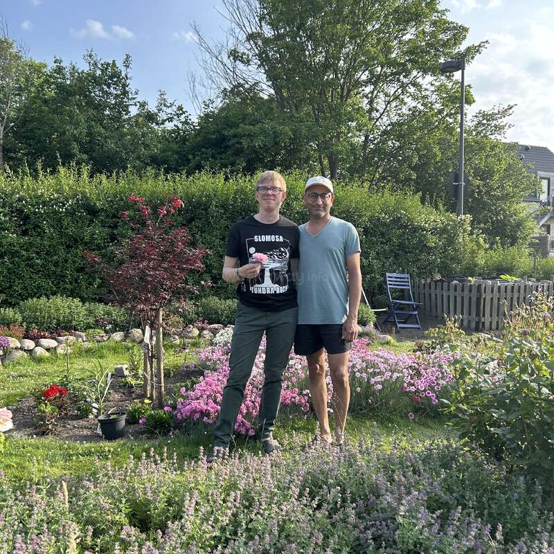 Deux hommes se tiennent côte à côte dans un jardin coloré, regorgeant de fleurs et de verdure. Ils semblent heureux, profitant d'une journée ensoleillée, entourés de plantes et d'arbres luxuriants.